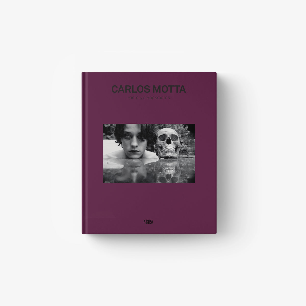 Deep purple hardcover book titled "Carlos Motta" with black-and-white photo of a man and a skull on the cover.