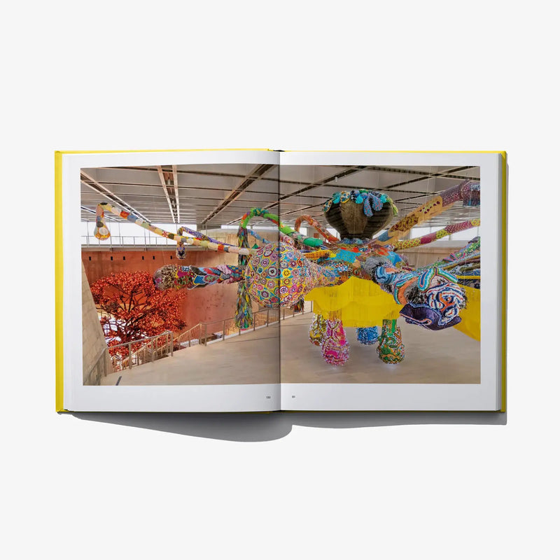 Open art book spread with photos of colorful textile figures inside a gallery.