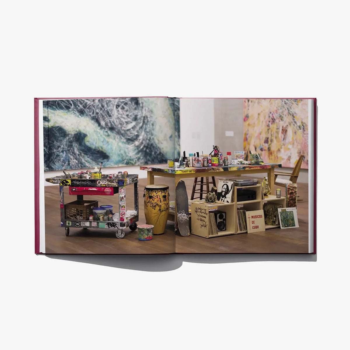 Open hardcover art book with studio display of paintings and prints on tables